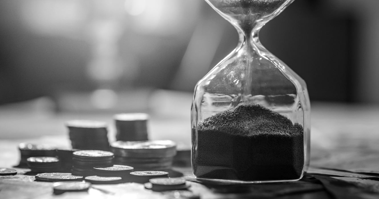 A black-and-white close up image of a hourglass with stacks of coins next to it