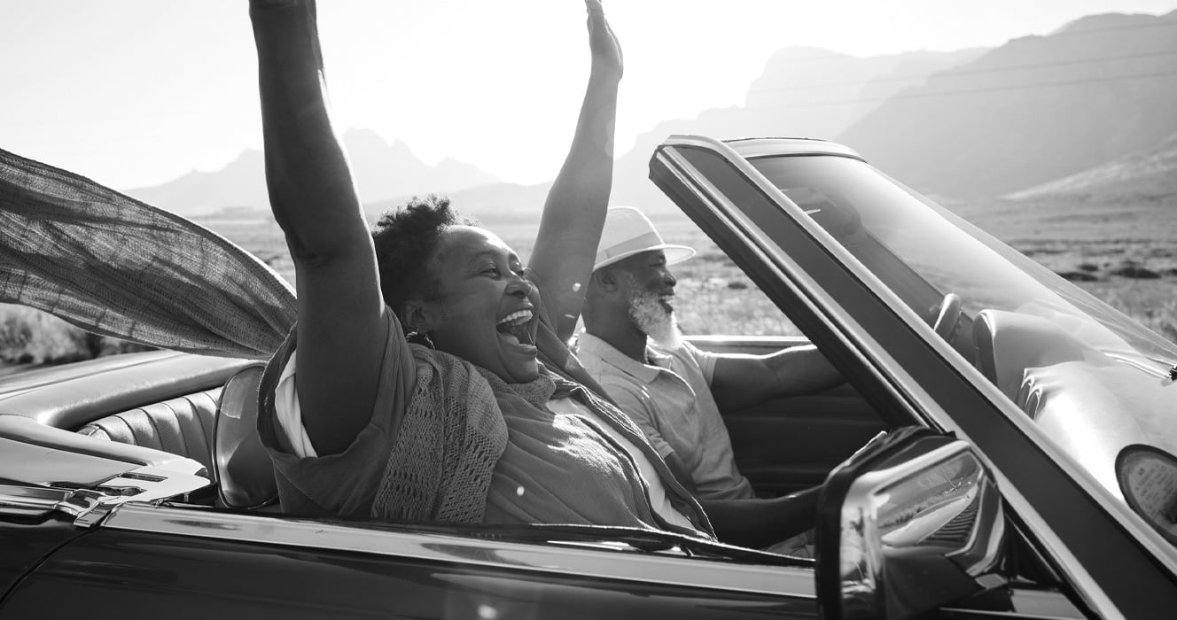 Black and white image of a couple riding in a convertible with the woman holding her hands up